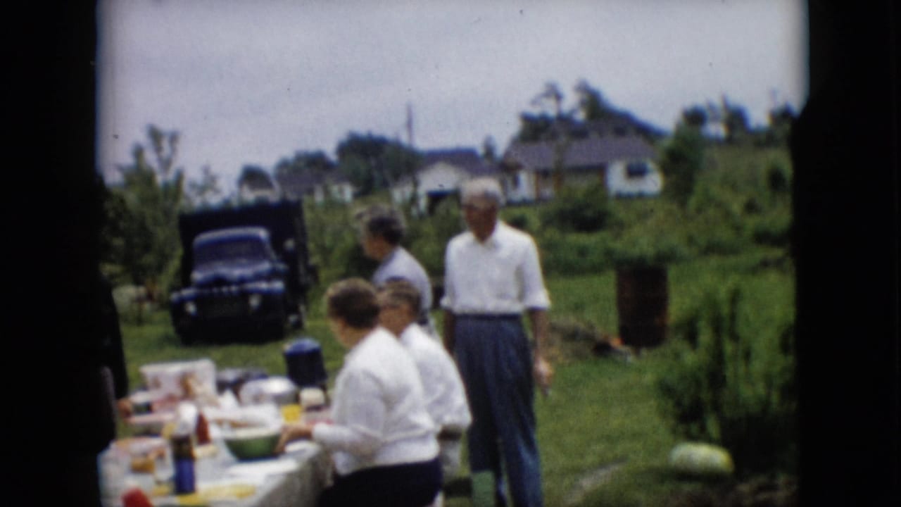 1957 Pennsylvania home-movie frame showing adults in 1950s clothing around a backyard meal table, with lawn, garden greenery, houses, and a dark parked vehicle behind them.
