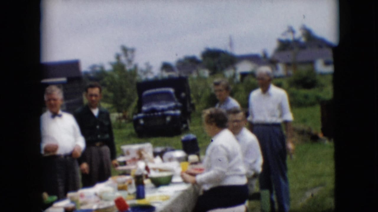Several adults stand and sit beside a covered outdoor table with dishes and a dark vehicle beyond the lawn.