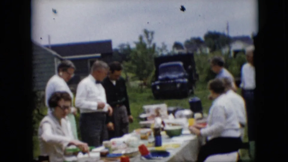1957 Pennsylvania home-movie frame showing adults in white shirts and dresses gathered around a backyard table during an outdoor meal, with a dark vehicle and houses behind them.
