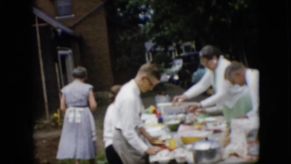 1957 Pennsylvania backyard meal frame showing adults serving food at a table near a building wall, with dishes, bowls, trees, and a parked vehicle partly visible in the background.