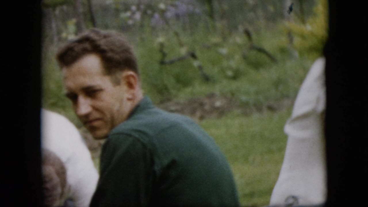 In a Pennsylvania 1957 home-movie frame, a man in a green shirt sits or crouches on a lawn and turns his head toward the camera while other adults are partly visible nearby.