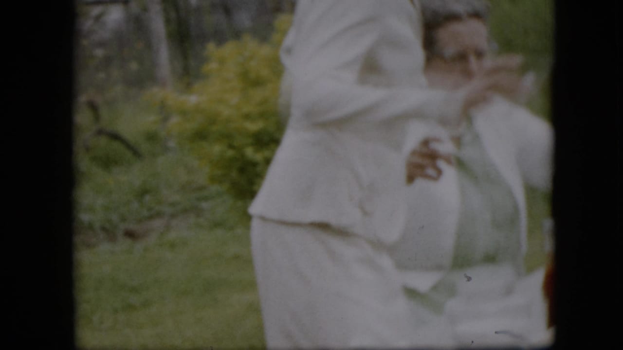 Adult in white stands in front of a seated woman during outdoor table preparation.