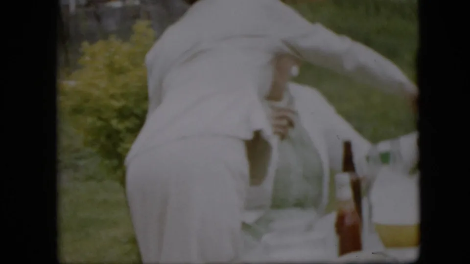 A white-clad adult bends near a table with bottles and a pitcher in a 1957 Pennsylvania home-movie frame from an outdoor dinner preparation scene.