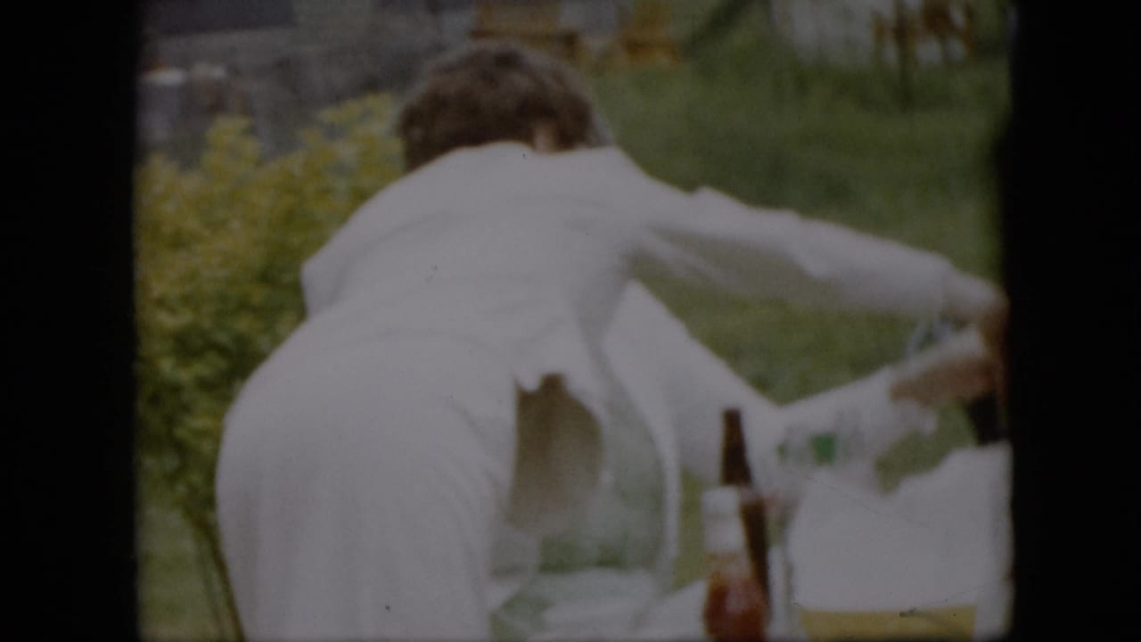 In 1957 Pennsylvania home-movie footage, a woman in a white dress or apron leans across an outdoor picnic table with bottles, cups, grass, and yellow flowers visible around her.