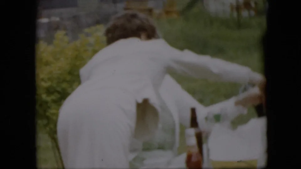 In 1957 Pennsylvania home-movie footage, a woman in a white dress or apron leans across an outdoor picnic table with bottles, cups, grass, and yellow flowers visible around her.