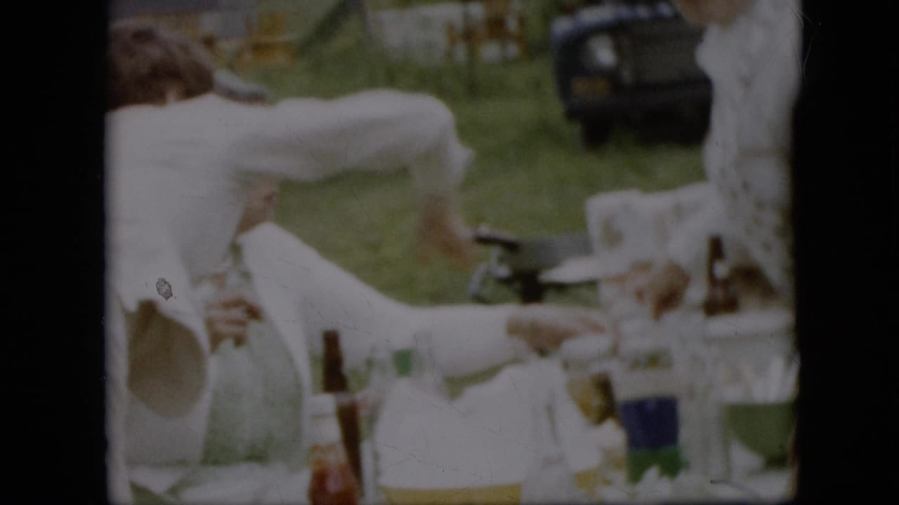 Women in white clothing gather around an outdoor picnic table in the 1957 Pennsylvania home movie, with bottles, cups, food containers, grass, and a dark vehicle in the background.