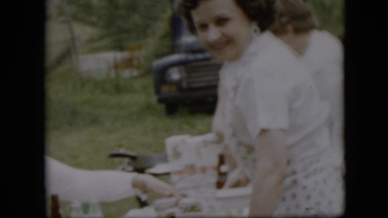 Smiling woman in a white top stands beside a picnic table with a dark truck behind her.