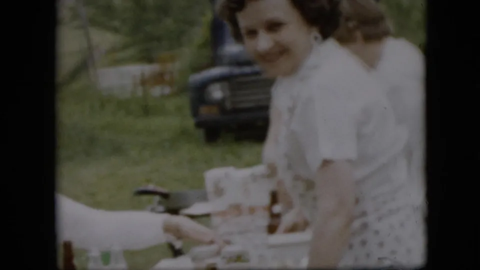 A smiling woman in white stands near the picnic table in this 1957 Pennsylvania home-movie frame, with bottles, cups, grass, trees, and a dark vintage truck behind her.