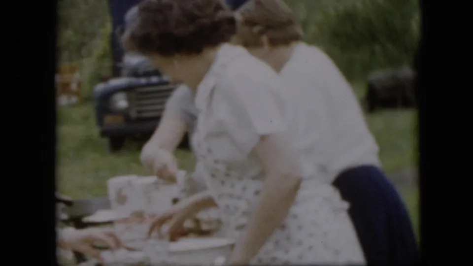 Two women in white clothing and aprons lean over an outdoor picnic table in the 1957 Pennsylvania footage, with bottles, cups, grass, and a dark truck visible behind them.