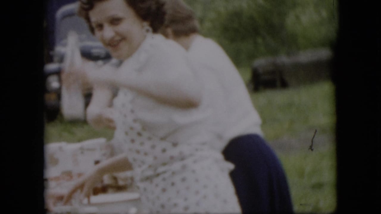 In 1957 Pennsylvania home-movie footage, a woman in a white blouse and dark skirt smiles while standing beside a picnic table with food containers on a grassy lawn.