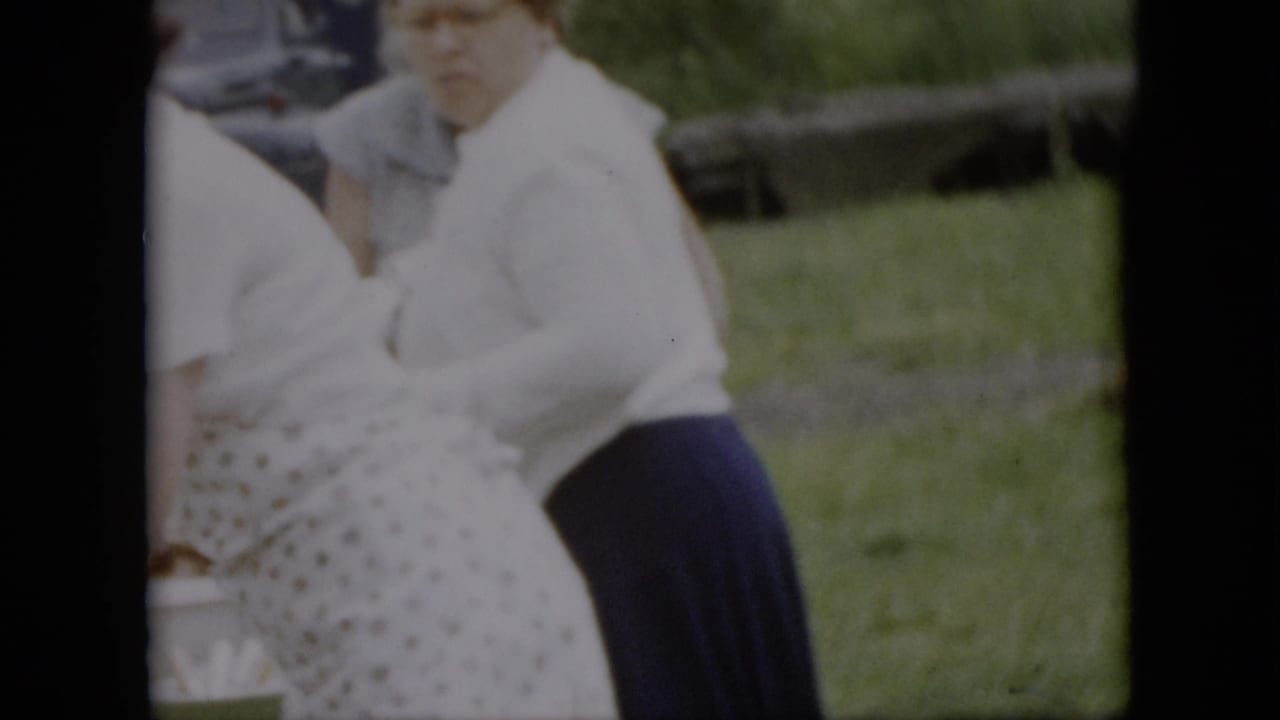 Two adult women stand near a picnic table in a grassy outdoor setting, one wearing a white blouse and dark skirt and another partly visible in a patterned apron.