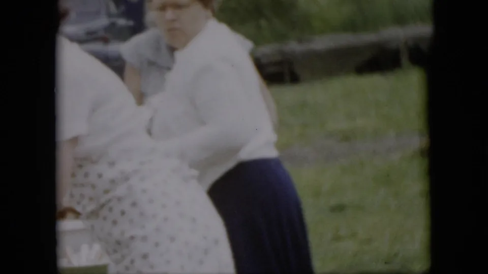 Two adult women stand near a picnic table in a grassy outdoor setting, one wearing a white blouse and dark skirt and another partly visible in a patterned apron.