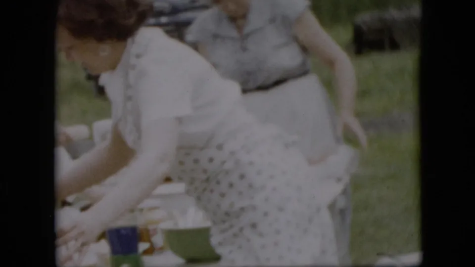 A woman in a patterned apron bends toward the picnic table as another adult woman stands behind her on the lawn, with picnic containers visible at the edge of the frame.
