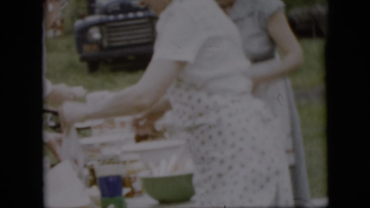 1957 Pennsylvania home-movie frame showing women in light dresses or aprons setting up a picnic table on grass, with a dark mid-century vehicle in the background.