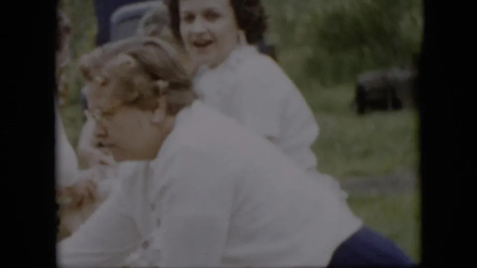 1957 Pennsylvania home-movie frame showing adults clustered around an outdoor picnic setup, including a woman facing camera and another person leaning forward in the foreground.