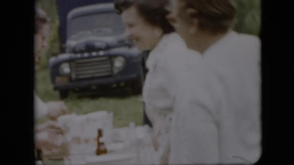 1957 Pennsylvania home-movie frame of two women beside an outdoor picnic table, with cups and dishes in the foreground and a dark truck parked behind them.