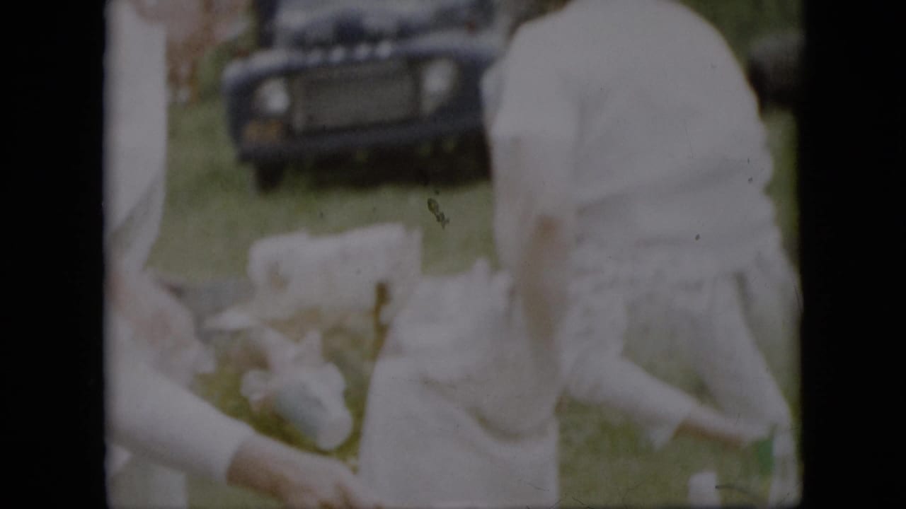 In a Pennsylvania 1957 home-movie frame, women in white dresses appear to prepare items at an outdoor picnic table while a dark older Ford is parked in the grassy yard behind them.