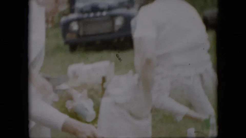 In a Pennsylvania 1957 home-movie frame, women in white dresses appear to prepare items at an outdoor picnic table while a dark older Ford is parked in the grassy yard behind them.