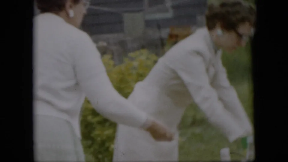 Two women in light dresses or sweaters stand in a Pennsylvania backyard garden, one reaching toward the table or another person during picnic setup in the supplied 1957 home-movie footage.