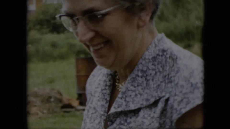 An older woman wearing glasses and a patterned blouse smiles in close view during the Pennsylvania backyard gathering, with lawn and garden objects behind her.
