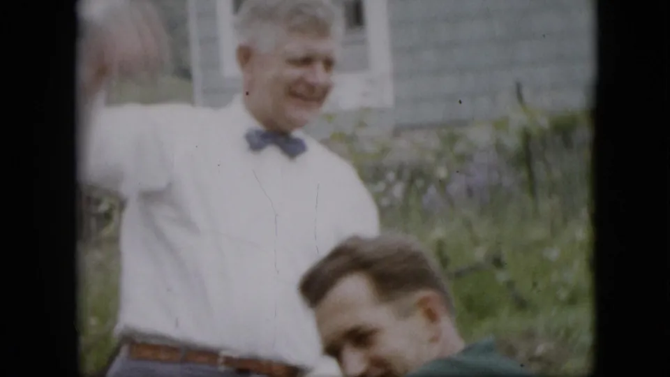 A man in a white shirt and bow tie stands in a garden yard while another man in a green shirt sits in the foreground, with house siding and plants in the background.