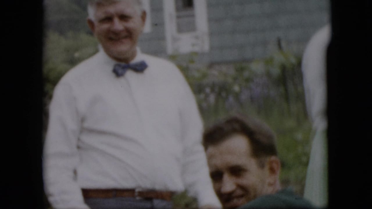 In this 1957 Pennsylvania home-movie frame, an older man in a white shirt and blue bow tie smiles in a grassy yard while another man is partly visible in front of him.