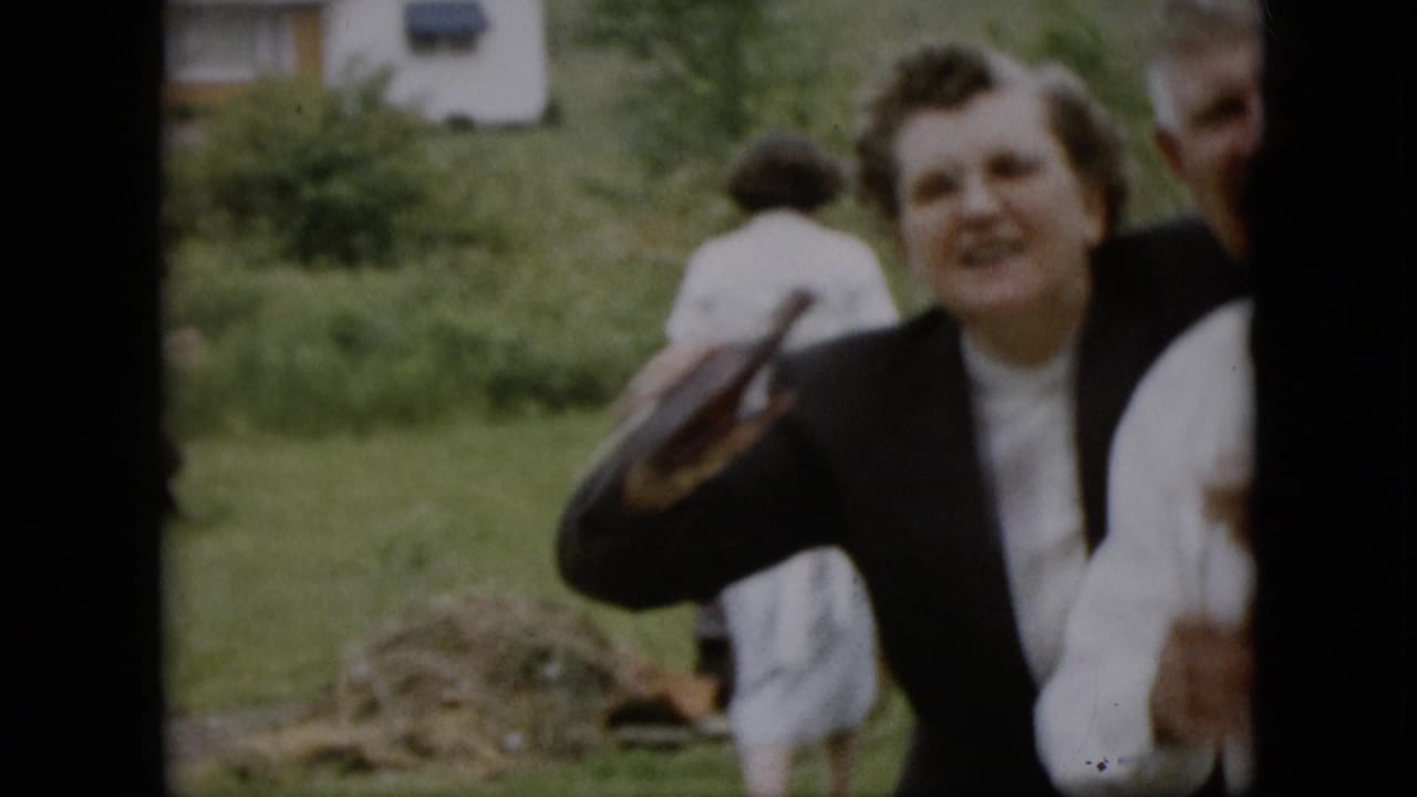 Woman in a dark jacket raises a bottle while laughing in a grassy yard with other adults nearby.