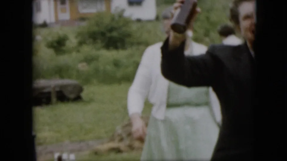 A woman in a white cardigan and pale skirt stands in the yard while the bottle-raising figure and another adult man are partly cropped at the right edge of the 1957 home-movie frame.