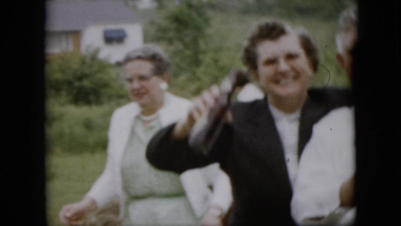 In 1957 Pennsylvania home-movie footage, adults gather in a grassy garden while a smiling woman in a dark jacket raises a bottle.