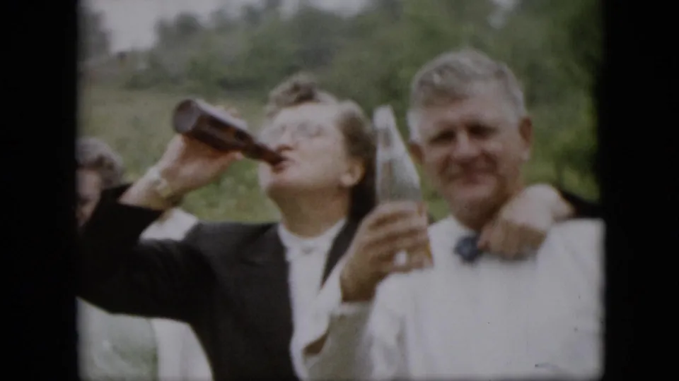 A woman in a dark jacket drinks from a bottle as a man in a white shirt and bow tie holds a glass during the 1957 Pennsylvania gathering.