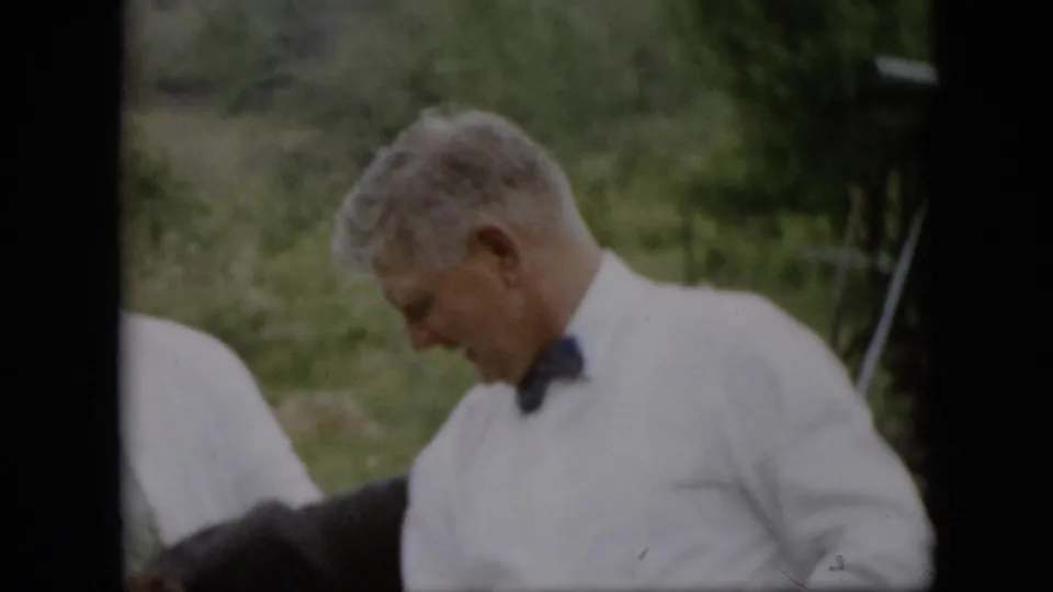 At the end of the Pennsylvania 1957 home-movie clip, a man in a white shirt and bow tie looks downward in the grassy outdoor setting.