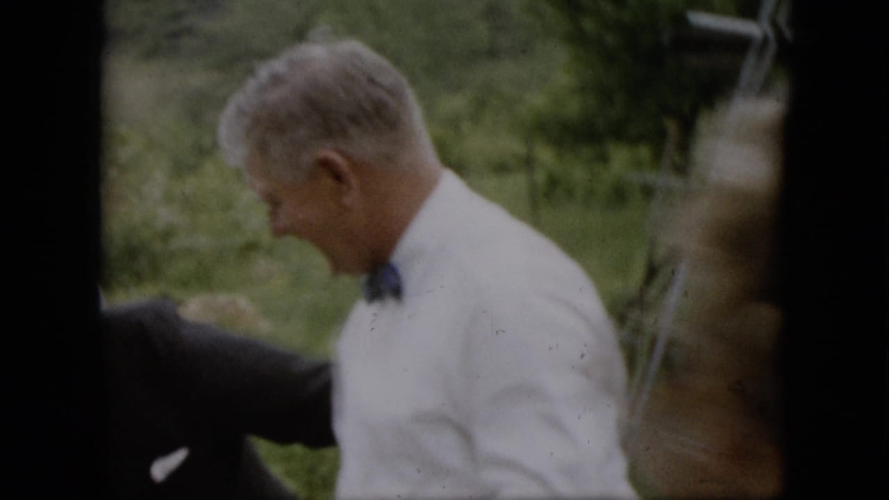 In supplied 1957 Pennsylvania home-movie footage, a man in a white shirt and dark tie is seen in profile in a grassy yard with other formally dressed adults partly visible.