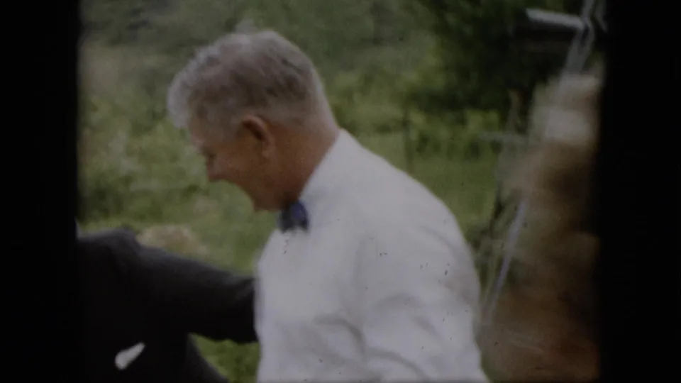 In supplied 1957 Pennsylvania home-movie footage, a man in a white shirt and dark tie is seen in profile in a grassy yard with other formally dressed adults partly visible.