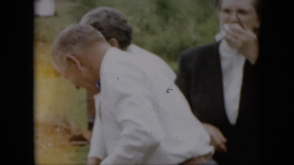 A man in a white shirt bends forward in the foreground as formally dressed adults behind him smile and gesture during the outdoor family celebration.