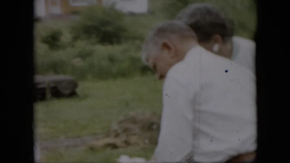 A man in a white shirt is seen from behind near a woman in a light cardigan, with grass and yard objects in the background of the 1957 Pennsylvania home movie.