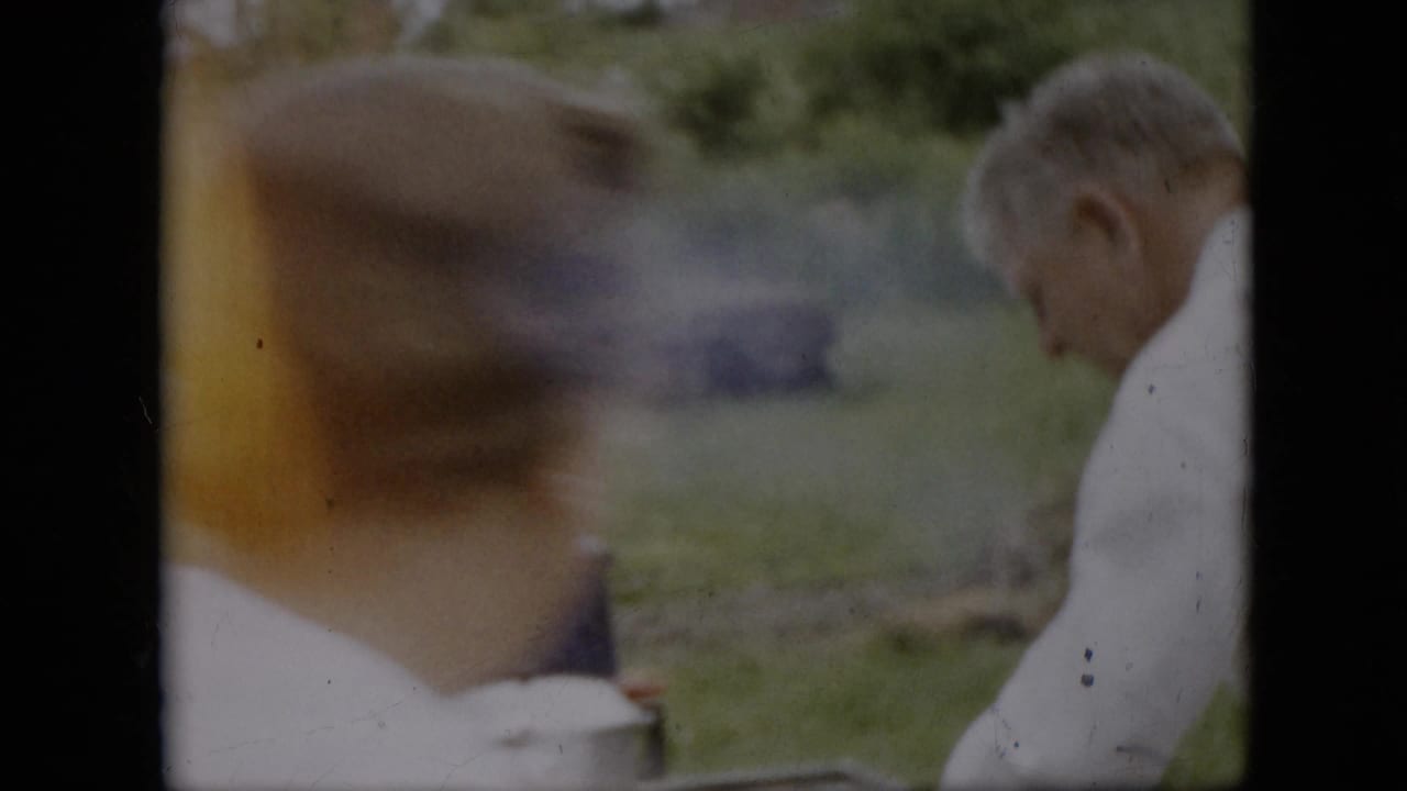 In this 1957 Pennsylvania 8mm home-movie frame, an older man in a white shirt appears to tend an outdoor cooking setup as a blurred person moves across the foreground.