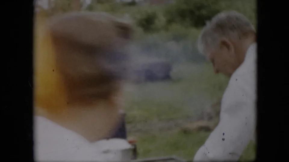 In this 1957 Pennsylvania 8mm home-movie frame, an older man in a white shirt appears to tend an outdoor cooking setup as a blurred person moves across the foreground.