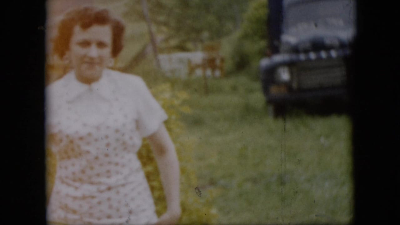 A woman in a white patterned dress smiles toward the camera in a grassy Pennsylvania yard, with trees, a small structure, and a dark older car visible in the background.