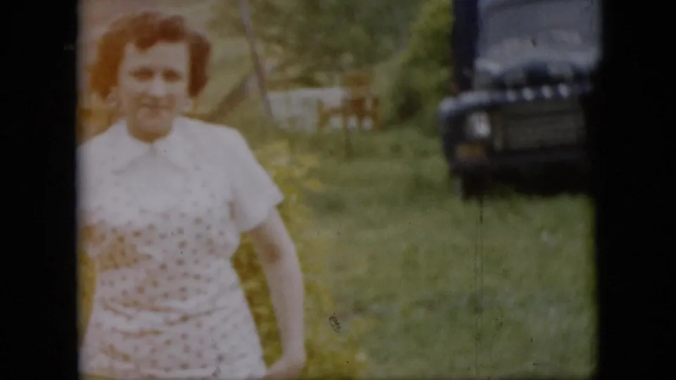 A woman in a white patterned dress smiles toward the camera in a grassy Pennsylvania yard, with trees, a small structure, and a dark older car visible in the background.