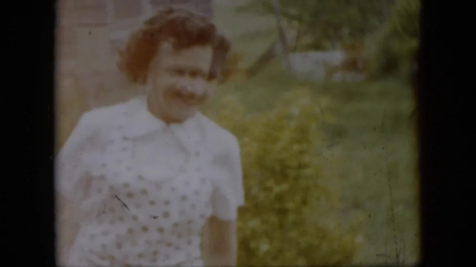 The home-movie frame shows the same woman in a white patterned dress standing outdoors near a building edge, grass, and garden-like greenery.