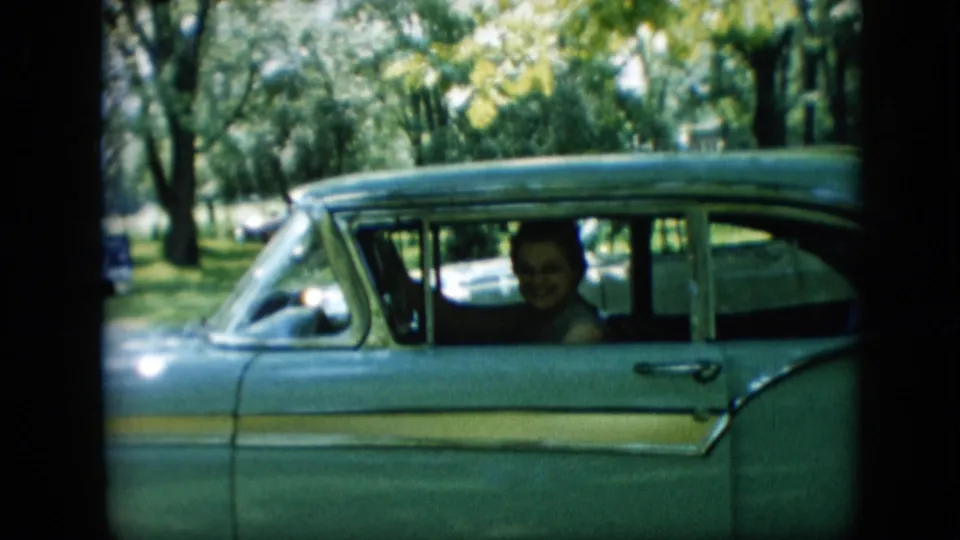 A woman smiles from the passenger seat of a green 1950s automobile in a sunlit Pennsylvania outdoor setting, captured on 8mm home-movie film.