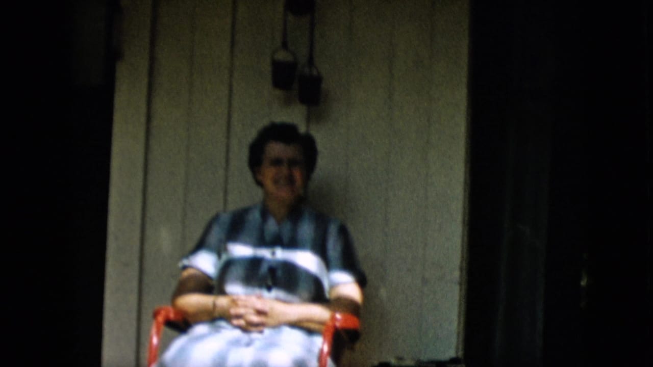In this 1957 Pennsylvania home-movie frame, an adult woman sits in a red chair on a shaded porch against light painted siding.