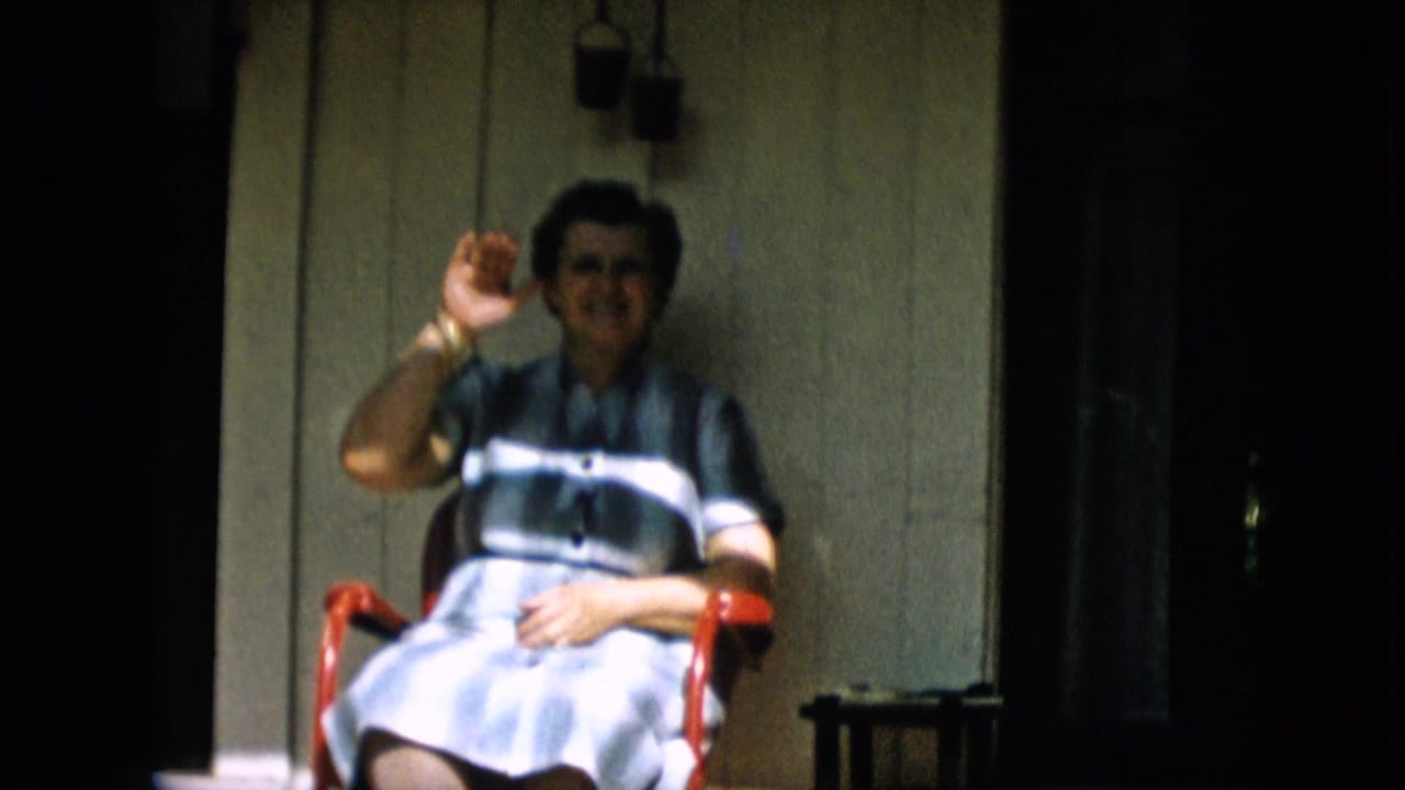 Woman seated on a porch raises one hand toward the camera.