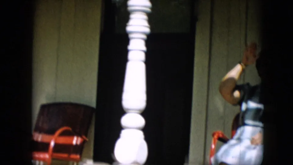 A white porch post cuts through the frame as the woman remains seated in the red chair, suggesting handheld filming from a passing or shifting viewpoint.