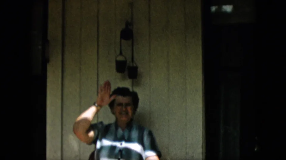 The adult woman sits on the shaded porch and waves toward the camera, framed by painted siding, hanging objects, and dark porch openings.