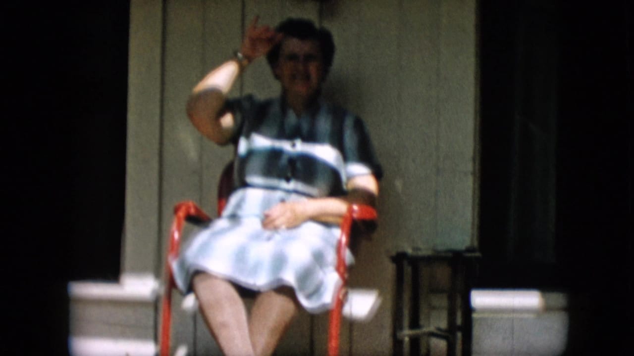In 1957 Pennsylvania home-movie footage, an older woman wearing spectacles and a blue-and-white striped dress sits on a red porch chair and lifts a hand toward the camera.