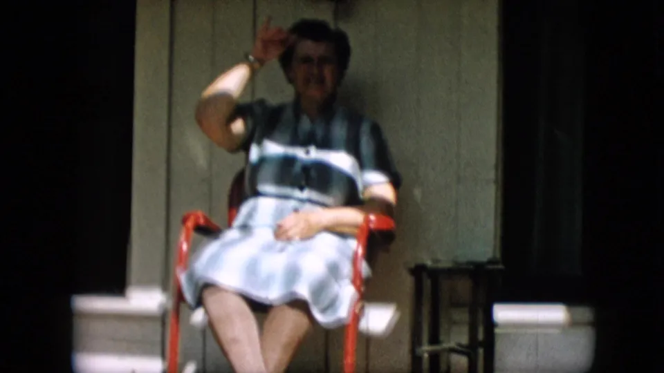 In 1957 Pennsylvania home-movie footage, an older woman wearing spectacles and a blue-and-white striped dress sits on a red porch chair and lifts a hand toward the camera.
