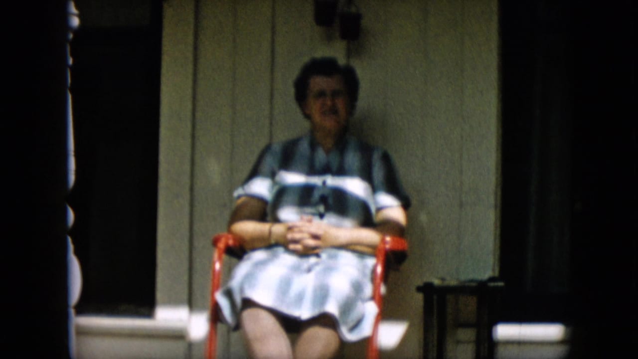 The woman sits facing the camera on a red chair, hands clasped in her lap, with the wooden porch wall, dark window, and small side table visible around her.
