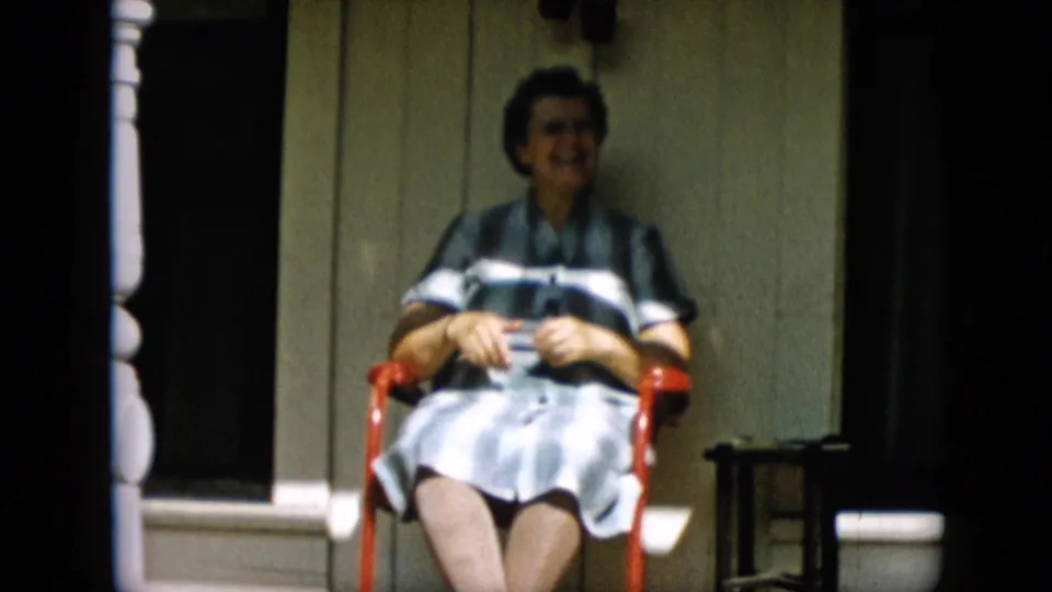 The woman remains seated on the red porch chair, her striped dress and spectacles visible as she looks toward the camera near a dark window and small table.