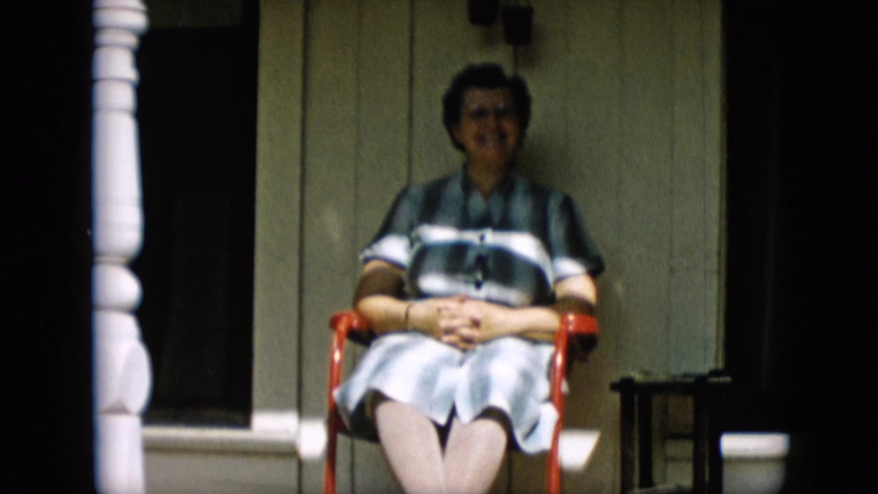 An older woman in a striped dress sits with hands clasped in a red chair on a porch, consistent with the 1957 Pennsylvania home-movie metadata.